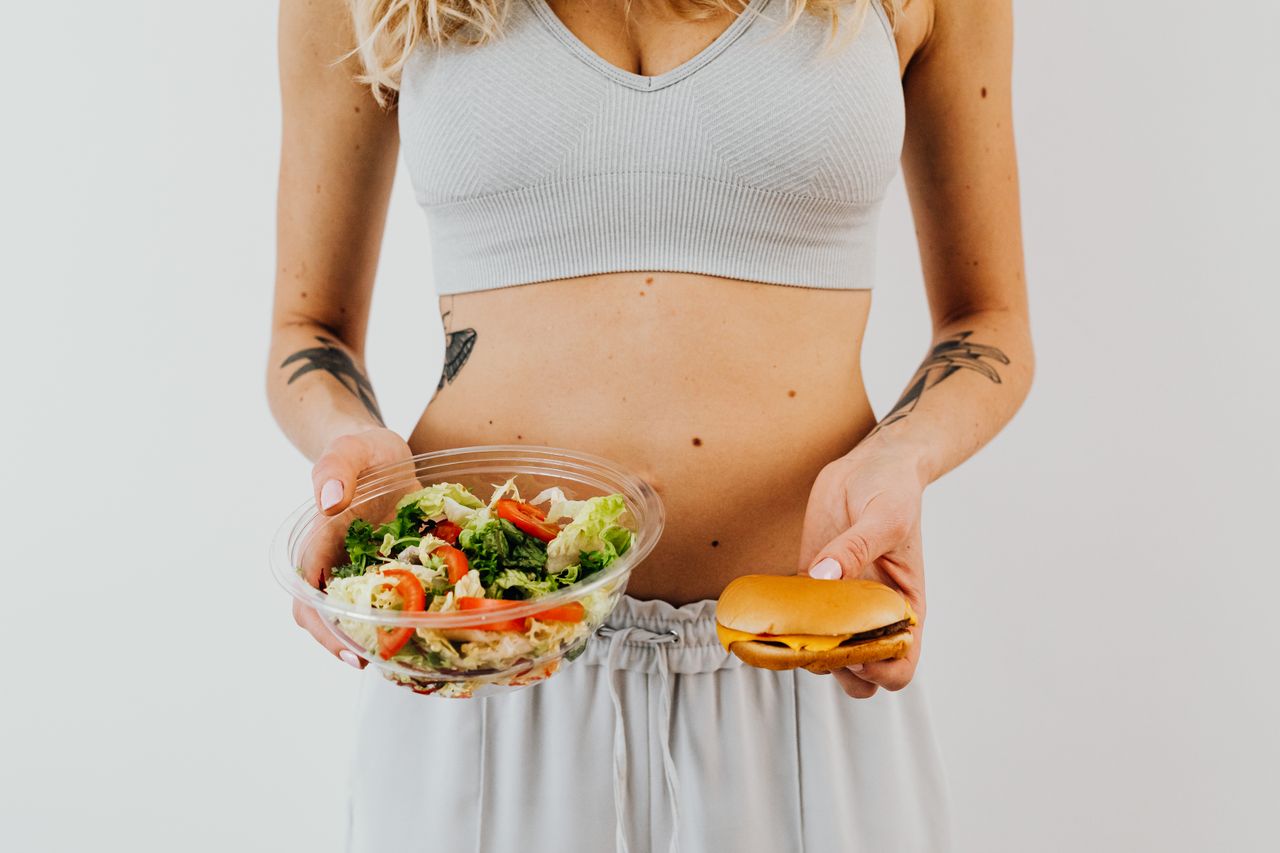 services-04 A woman in a sports bra holding a salad and cheeseburger, representing healthy vs junk food choices.